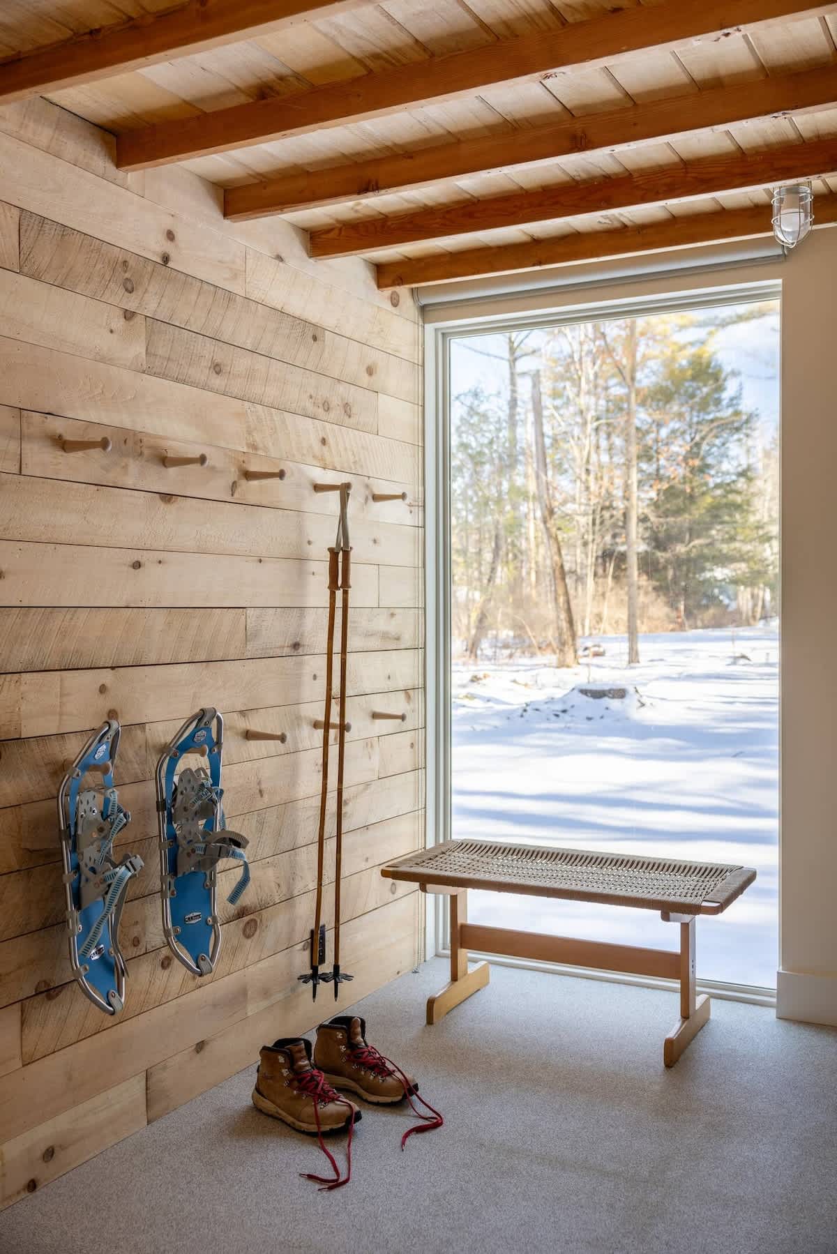 A rustic mudroom features a wooden plank wall with pegs holding snowshoes and ski poles, a woven bench, and a large window offering a view of the snowy outdoors.