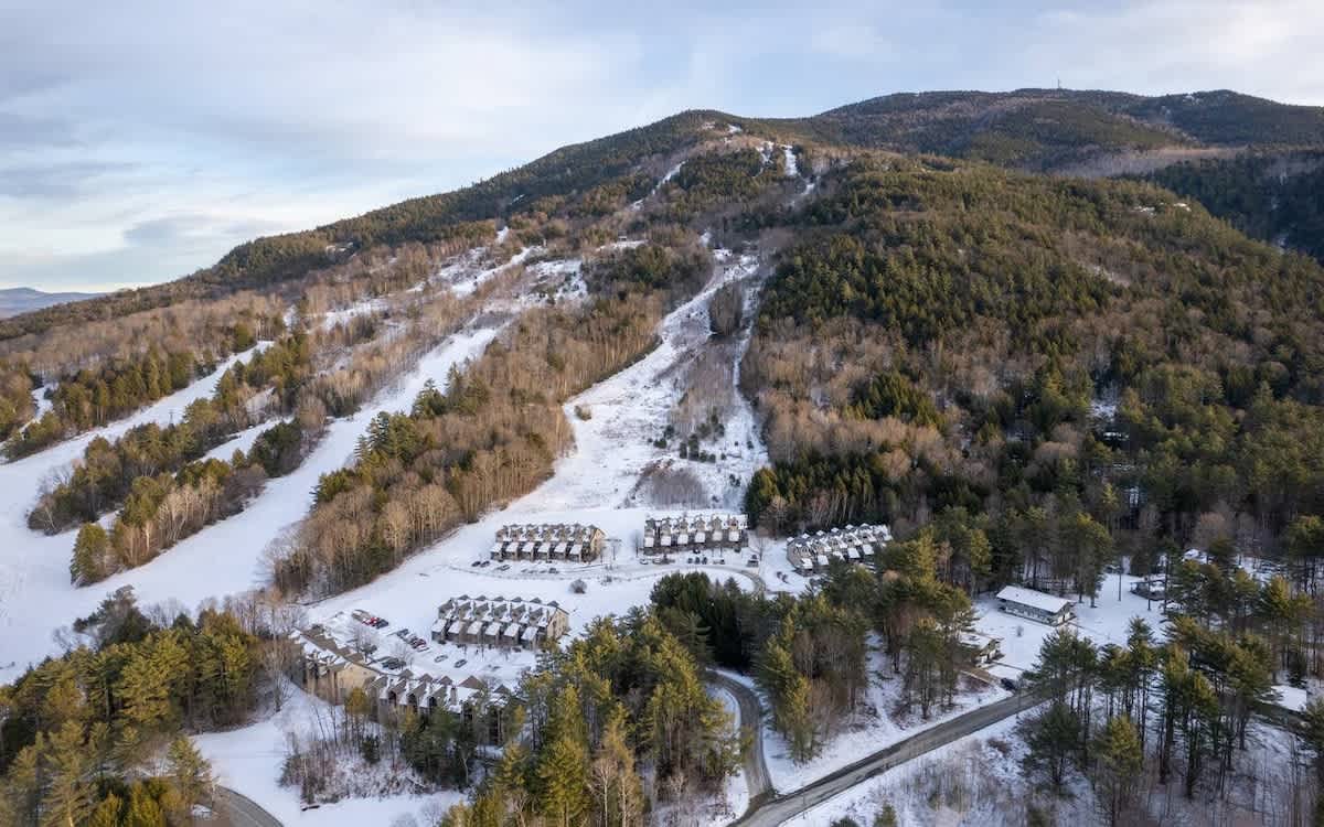 An expansive aerial view of a snow-covered mountain landscape with ski slopes and clusters of vacation rental properties nestled among evergreen and deciduous trees. A winding road is visible, indicating a winter resort setting.