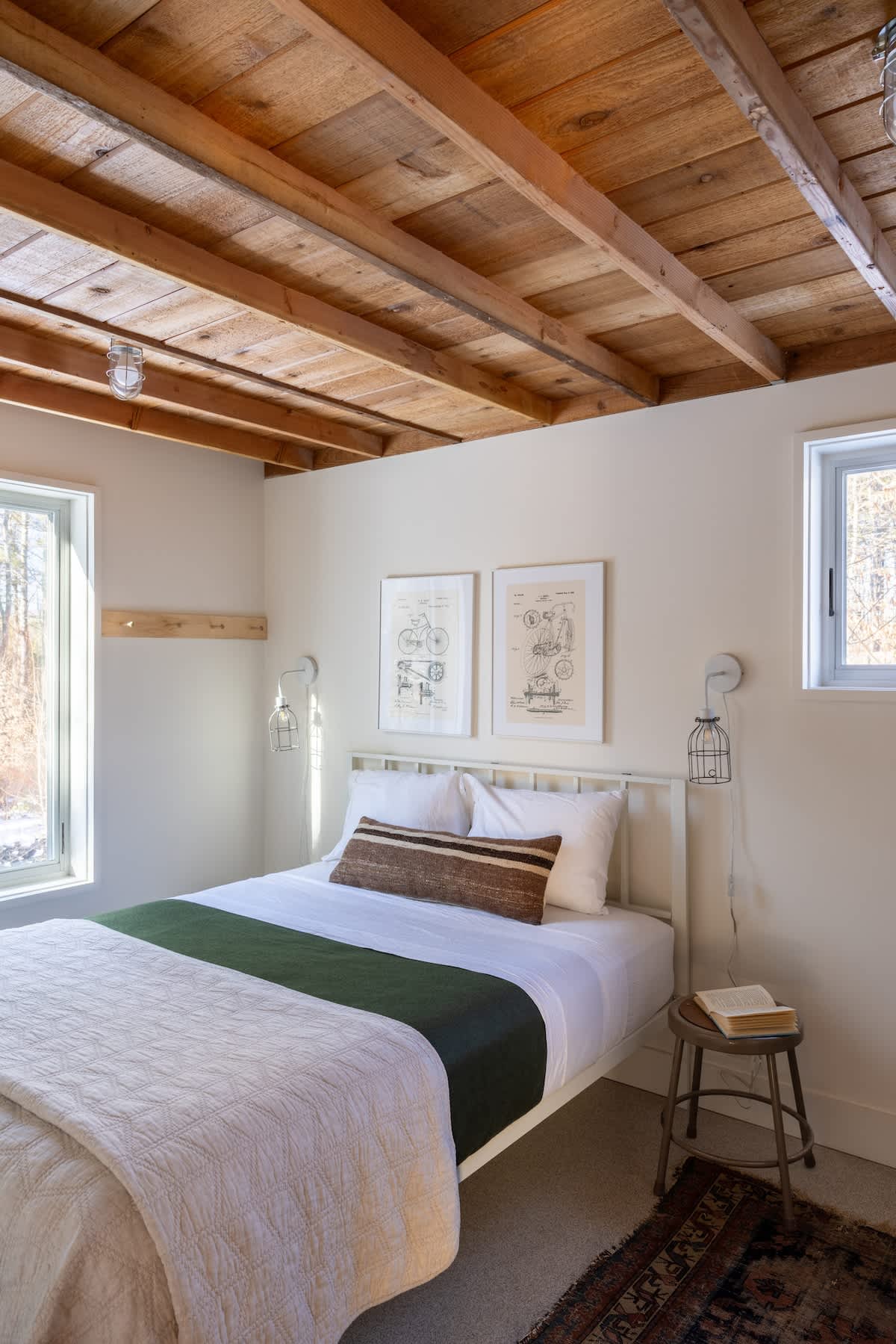 A bright bedroom features a white metal bed frame with white bedding and a green throw, flanked by two windows and industrial-style wall sconces. The ceiling is covered with rustic wooden planks, and two framed patent prints hang above the bed.