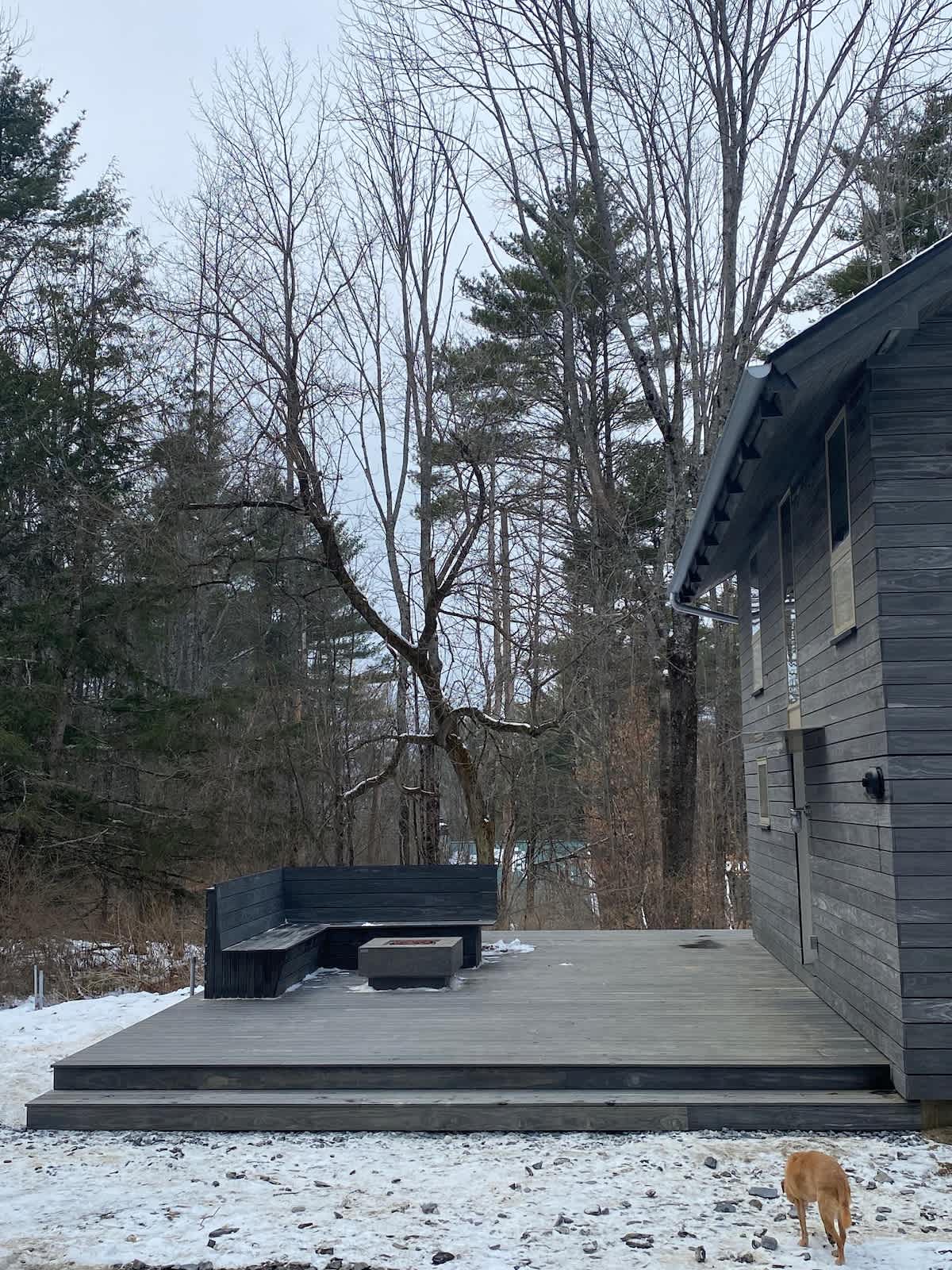 A modern dark grey wooden deck features a built-in L-shaped bench and a square fire pit, set against a snowy landscape with a dark-sided house and bare trees in the background. A light brown dog walks in the foreground.