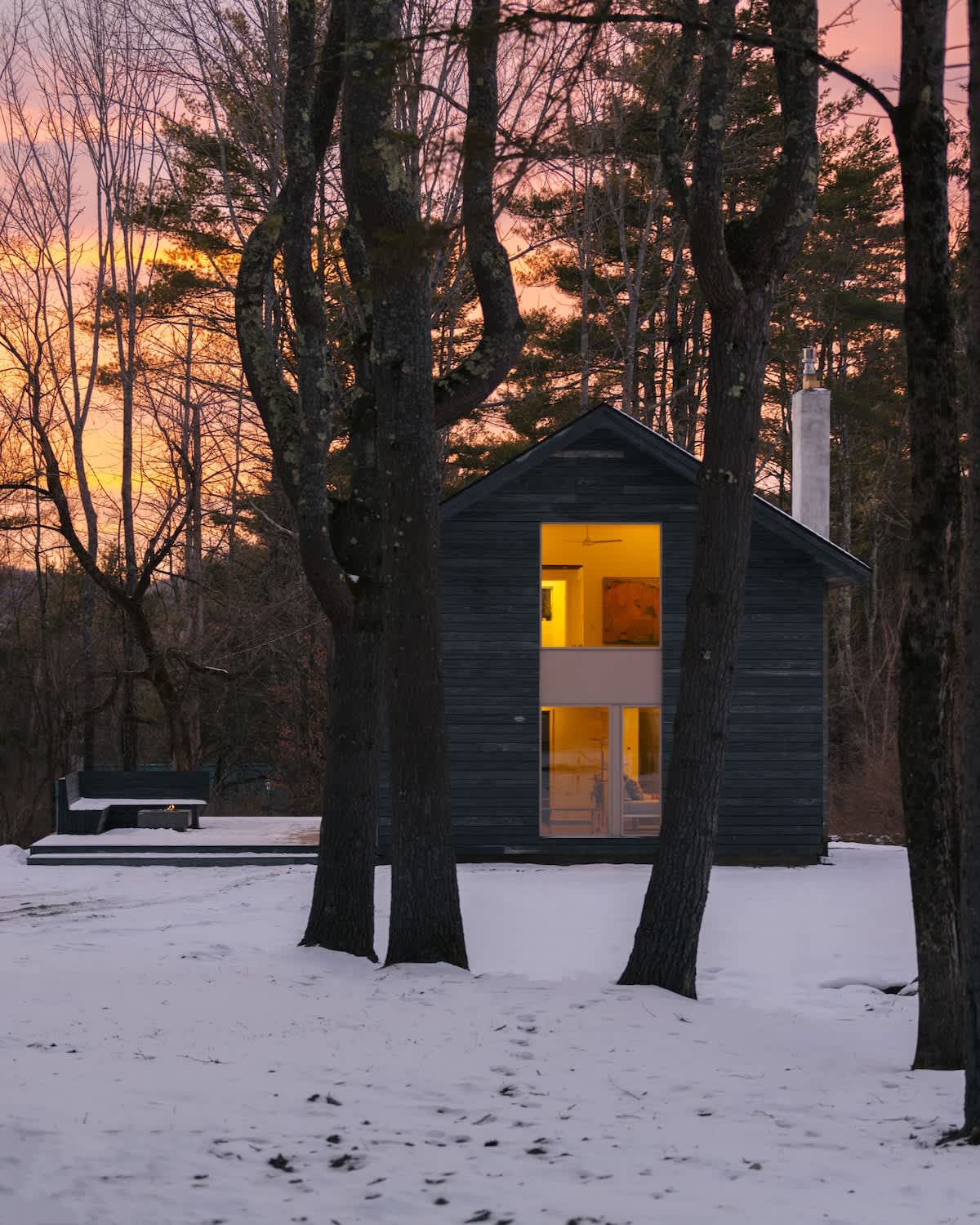 The front facade of the dark grey modern house, with its large windows glowing warmly, is framed by bare trees against a vibrant orange and pink sunset sky, with snow covering the ground.