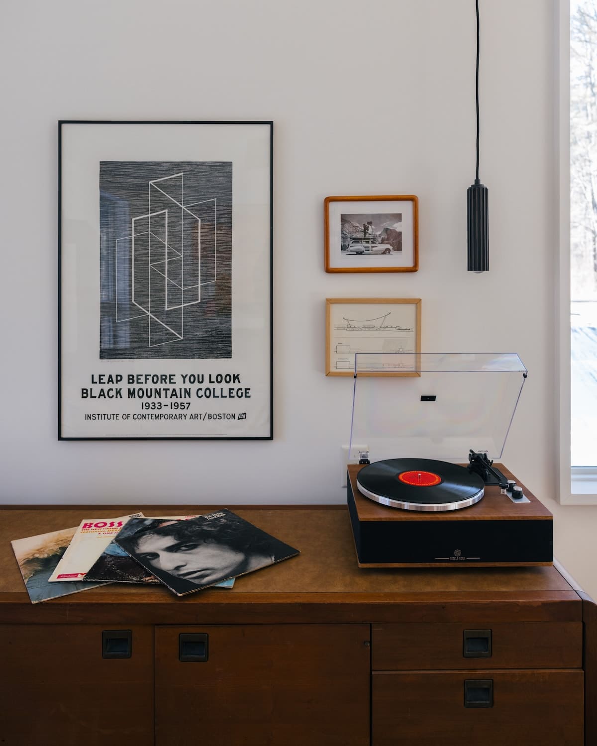 A stylish corner features a wooden credenza with a turntable and vinyl records, beneath a large framed geometric art print and smaller framed pictures on a white wall. A modern black pendant light hangs from the ceiling.