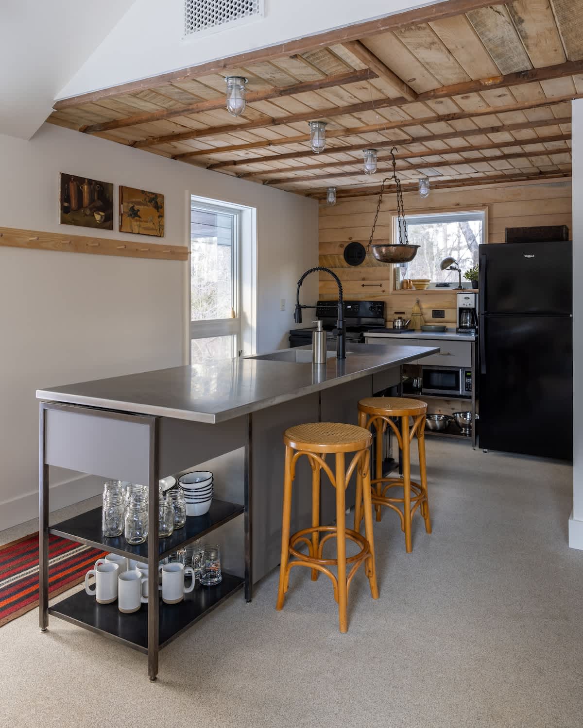A modern kitchen features a large island with a grey countertop and two rattan bar stools, black appliances including a refrigerator, and a rustic wooden plank ceiling with exposed beams.