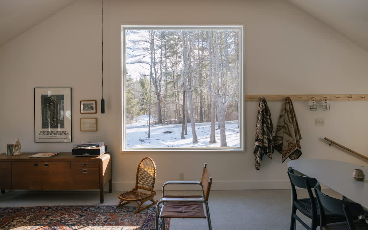 A spacious living area with a large window overlooking a snowy forest, featuring a wooden sideboard with a record player, a patterned rug, and various seating options.