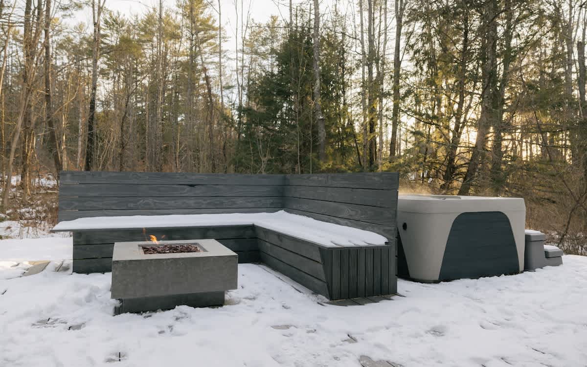 An outdoor deck area covered in snow, featuring a modern hot tub, built-in L-shaped seating, and a concrete fire pit with a small flame, all surrounded by a dense forest.