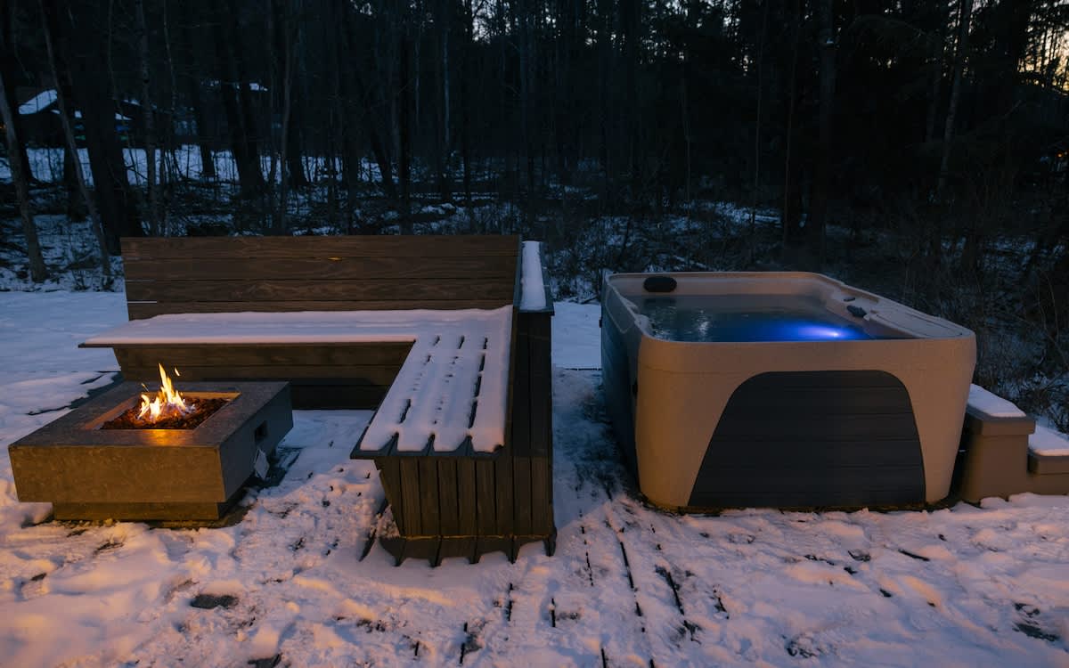 A bubbling hot tub with blue interior lighting sits on a snowy deck next to a built-in bench and a square fire pit with a flickering flame, surrounded by dark trees at night.