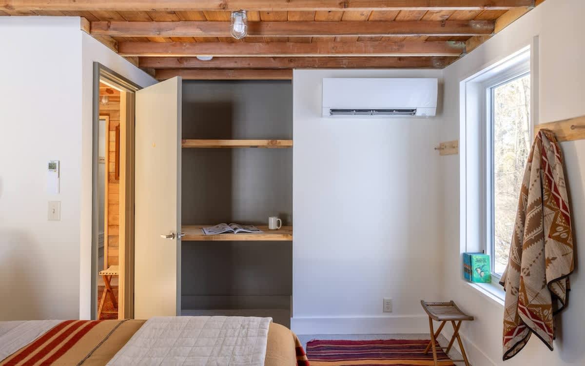 A functional corner of a bedroom features a built-in wooden shelf and desk area within an open closet, next to a window, with a rustic wooden ceiling and white walls. An air conditioning unit is mounted high on the wall.