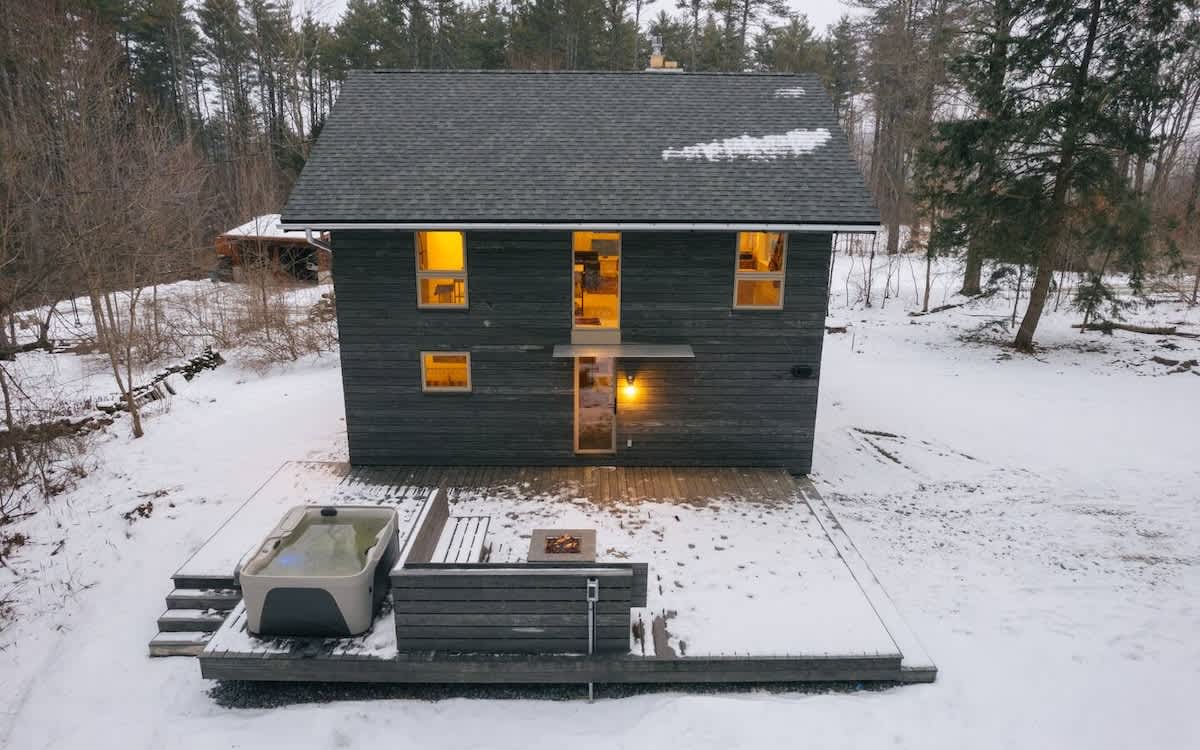 An aerial view of a modern dark-sided house with a gray shingle roof, surrounded by snow-covered ground and bare trees. A wooden deck extends from the back, featuring a hot tub and a fire pit area, with warm lights glowing from inside the house.