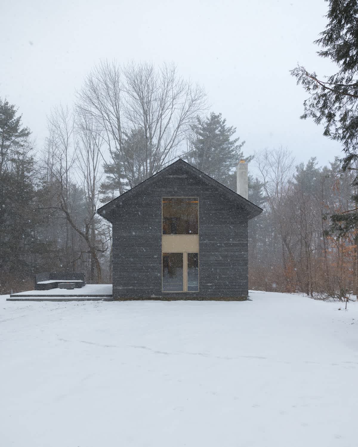The minimalist dark grey house stands starkly in a snowy landscape, with large front windows reflecting the overcast sky, as snowflakes fall gently around it and bare trees line the background.