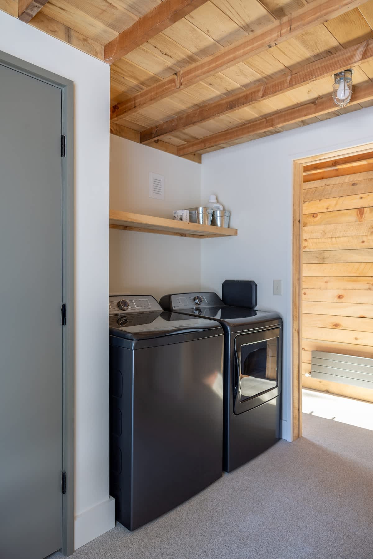 A functional laundry area features a dark gray washer and dryer set, with a wooden shelf above holding supplies, all beneath a rustic wooden plank ceiling.