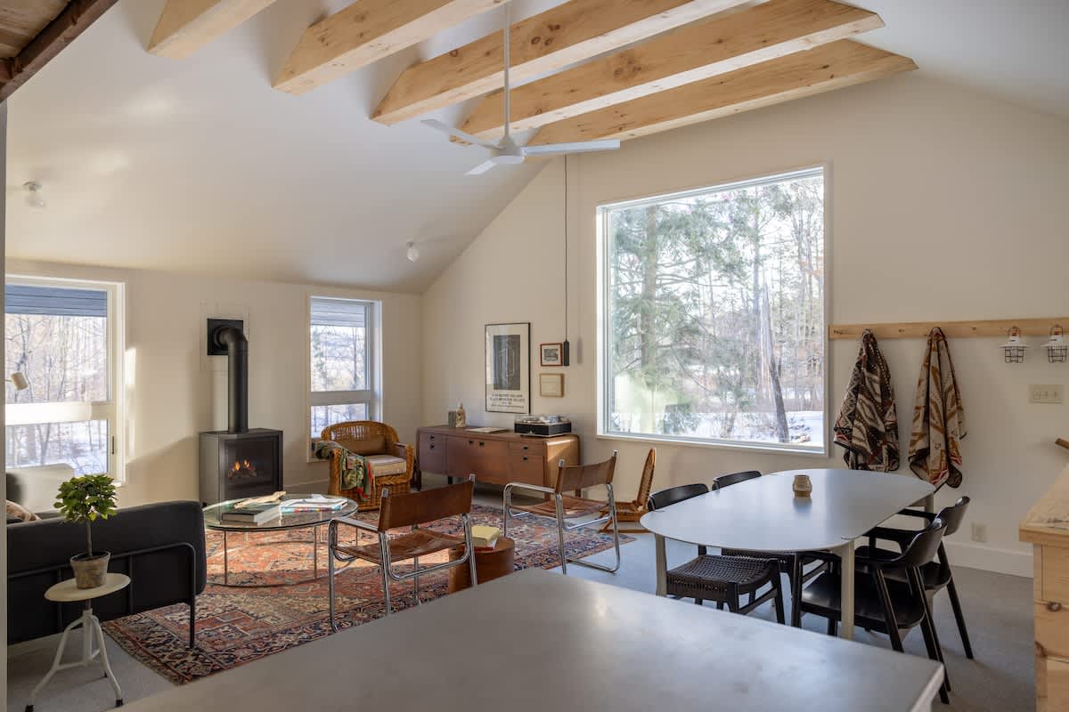 A bright, open-concept living and dining area with a vaulted ceiling and exposed wooden beams, featuring a dark sofa, a wood-burning stove, and a dining table with black chairs, all overlooking a snowy forest through large windows.