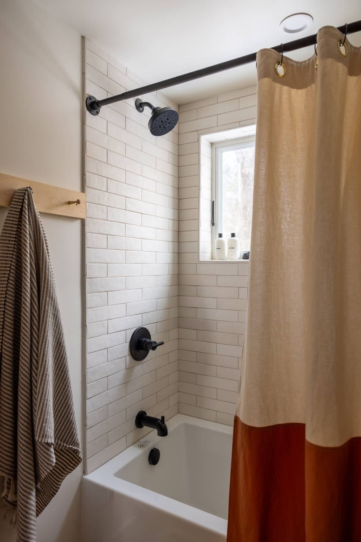 A modern bathroom featuring white subway tile walls in the shower area, a white bathtub, and matte black fixtures. A two-tone shower curtain in beige and rust orange hangs from a black industrial-style rod, with a wooden peg rack and striped towel visible on the left.