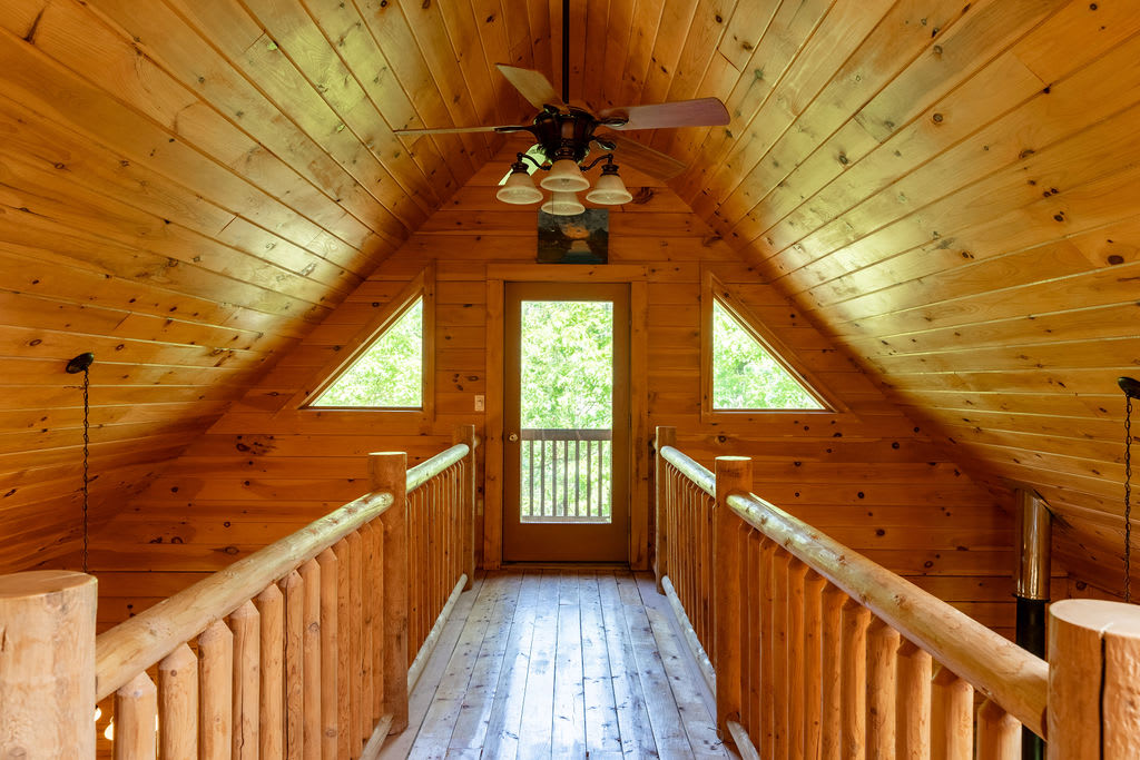 An upstairs landing with a wooden floor and sloped wood-paneled ceiling, featuring a door leading to a balcony flanked by two triangular windows, and log railings.