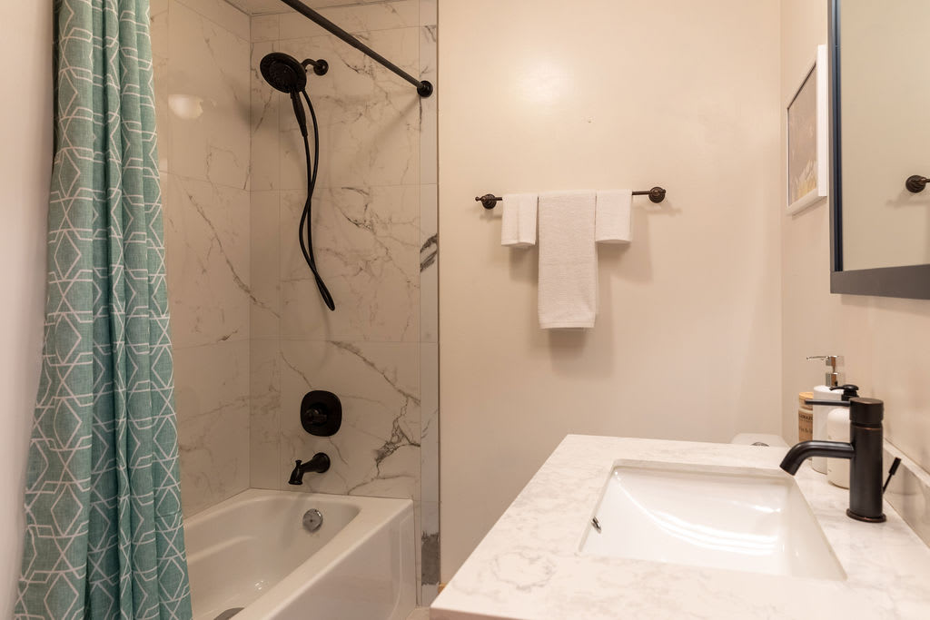 Modern bathroom with a white bathtub and shower featuring white marble-patterned tiles and a black shower head. A teal geometric shower curtain hangs to the left.