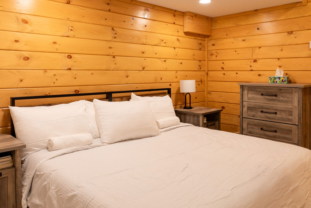 Bedroom with light wood paneled walls and a queen-sized bed with white bedding. A nightstand with a lamp and a dresser with a tissue box are visible.