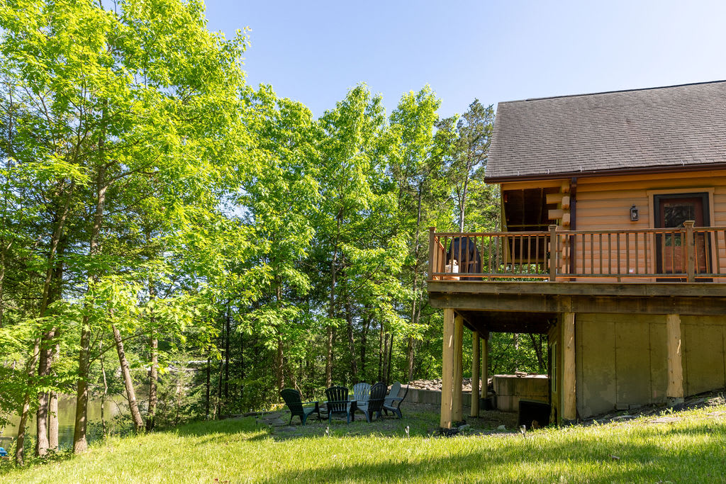 The back of the wooden cabin with a raised deck overlooking a grassy yard and a fire pit area, surrounded by vibrant green trees. Adirondack chairs are arranged around the fire pit.