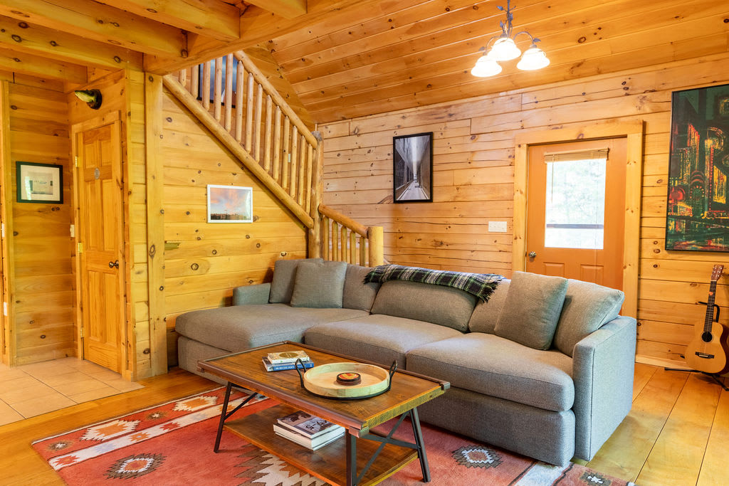 A rustic living room with light wood paneling and a staircase leading to an upper floor, featuring a large L-shaped sectional sofa with an area rug and a wooden coffee table. A guitar leans against the wall.