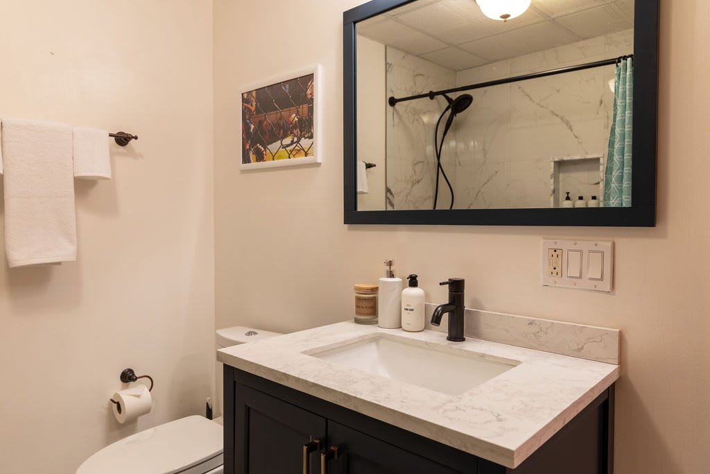 Modern bathroom with a dark vanity, white countertop, and rectangular sink. A large mirror with a black frame hangs above, reflecting the shower.