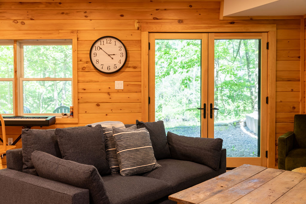 Living room with wood paneled walls, a dark gray sofa, and a large round wall clock. French doors open to an outdoor area with lush greenery.