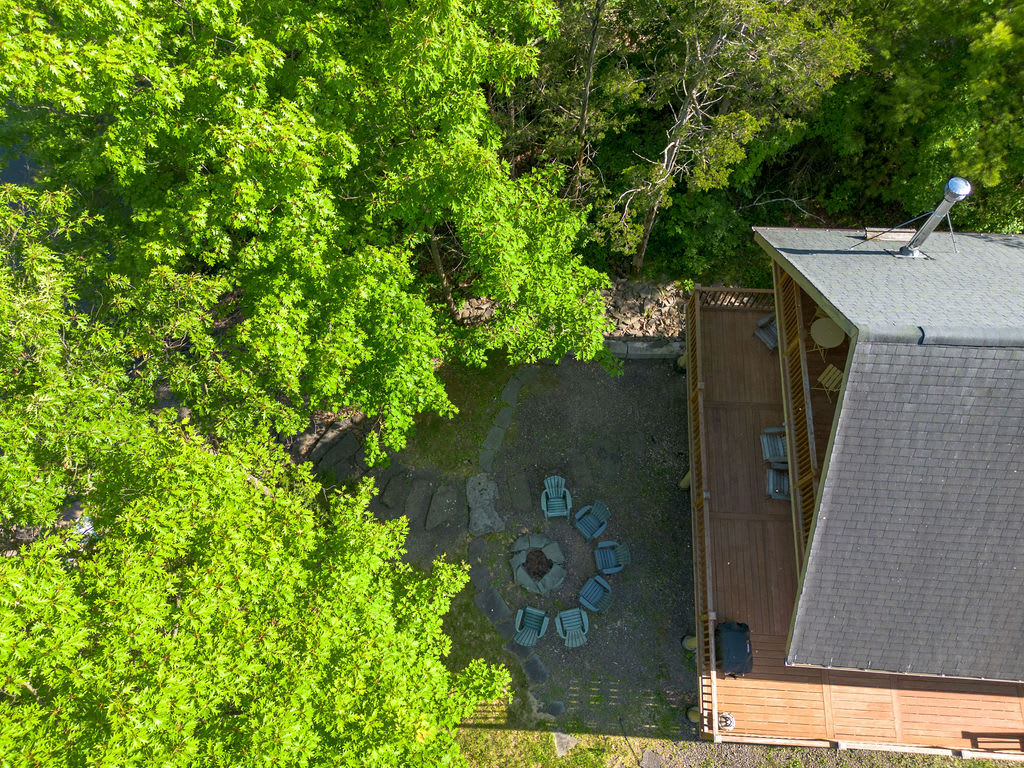 An overhead aerial view of the cabin's backyard, showing a large wooden deck, a gravel fire pit area with chairs, and lush green trees providing shade. The cabin's roof and chimney are visible.