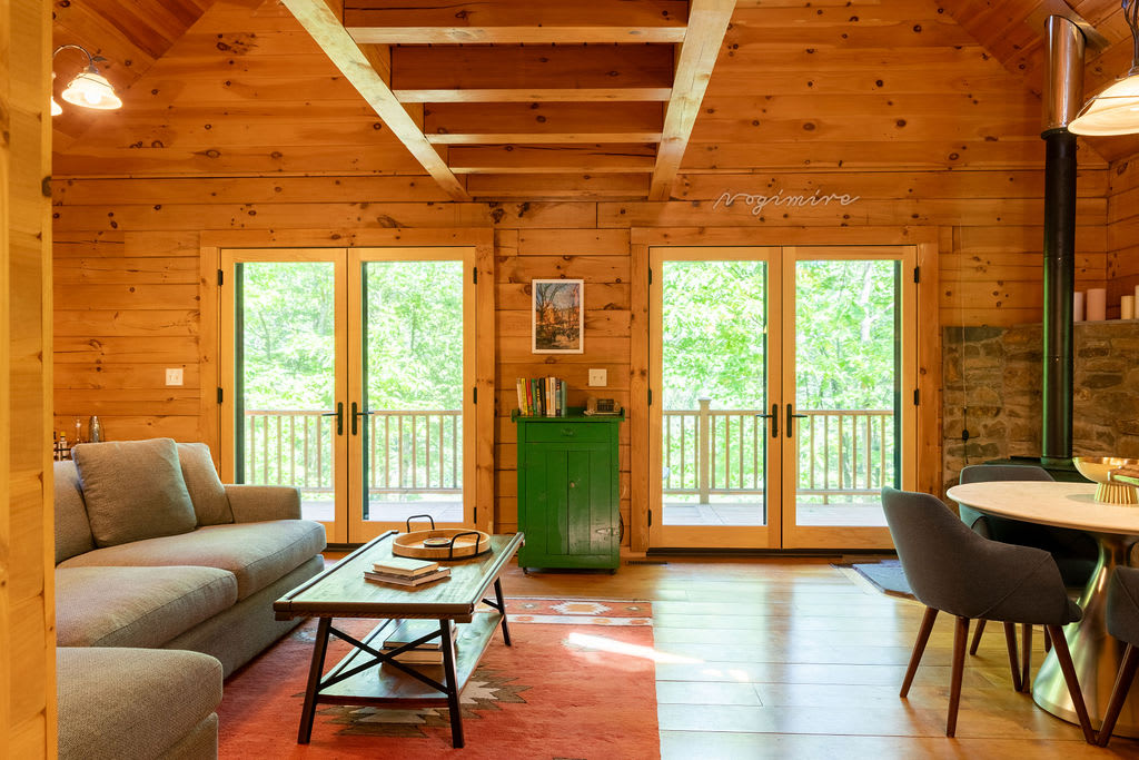 A bright living room with warm wood-paneled walls and ceiling, featuring a grey sectional sofa, a rustic wooden coffee table on a patterned rug, and two sets of French doors opening to lush greenery.