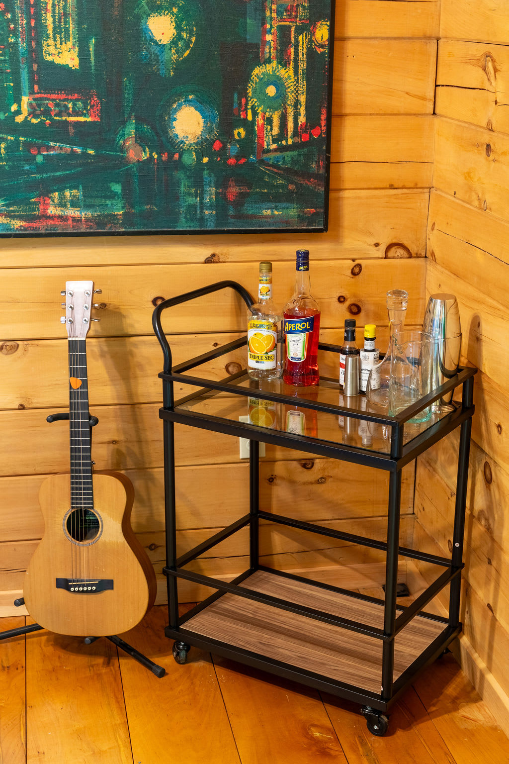 A cozy corner featuring a black metal bar cart stocked with bottles and glassware, positioned next to an acoustic guitar on a stand, all against warm wood-paneled walls.