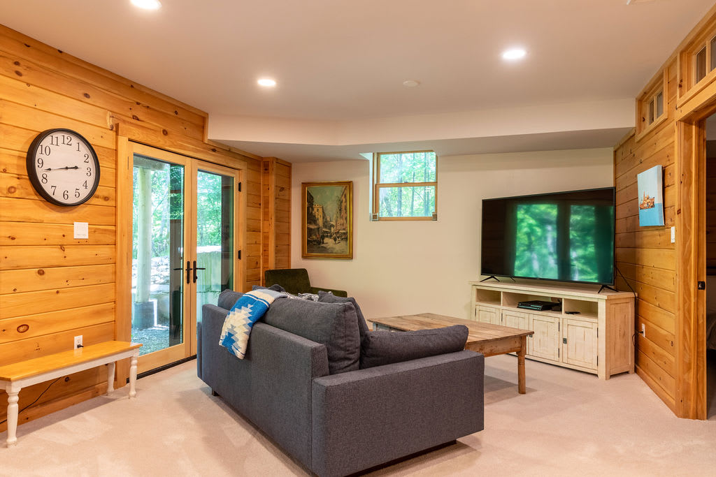 A cozy basement living area with light wood paneling on one wall and carpeted floors, featuring a large grey sofa, a wooden coffee table, and a flat-screen TV on a white console. French doors lead to the outside.