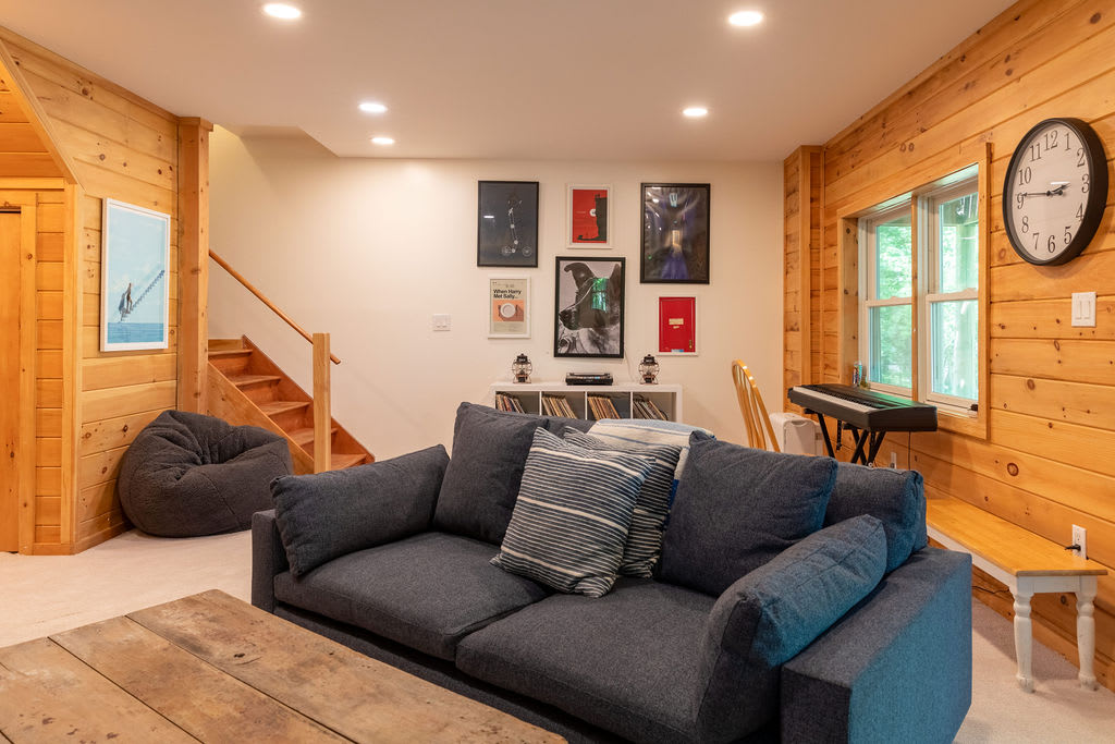 Cozy living room with light wood paneled walls, a large dark gray sofa with striped pillows, a rustic wooden coffee table, and a staircase leading upstairs. Art prints adorn a white wall.