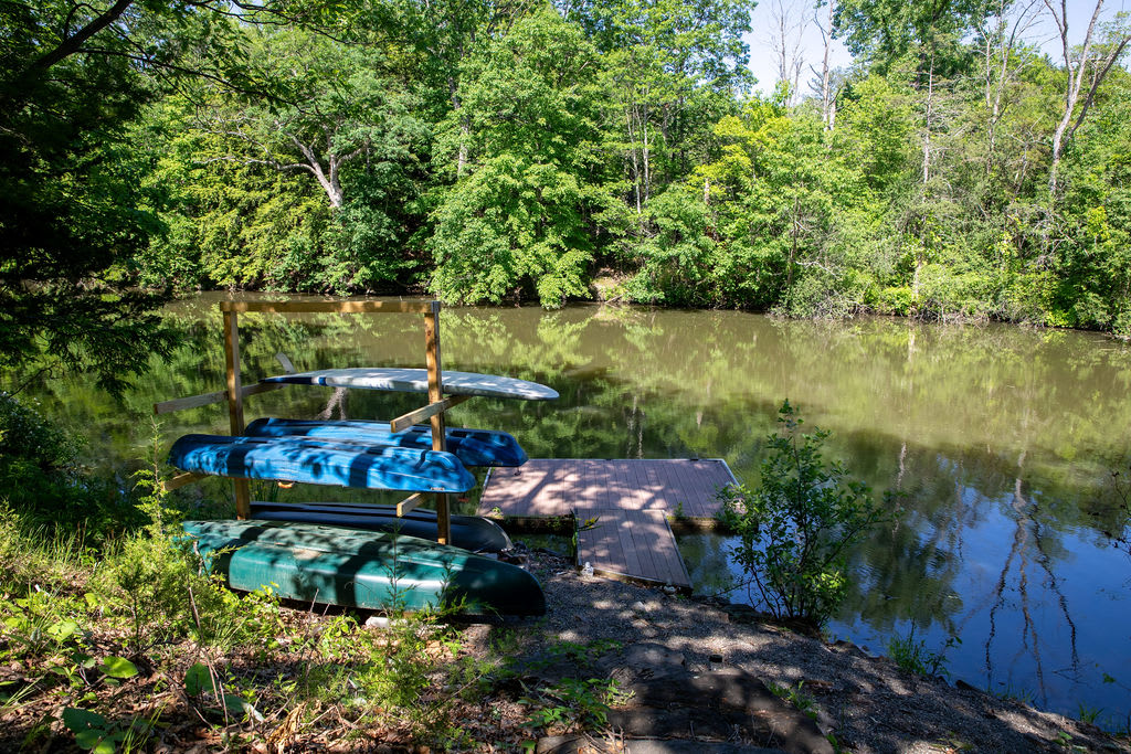 A serene lakefront scene with a wooden dock and a rack holding multiple kayaks and canoes, surrounded by dense green foliage. The calm water reflects the trees and sky.