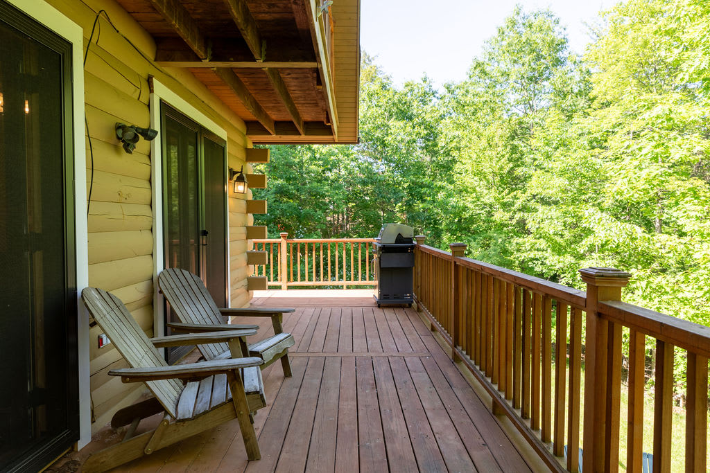 A rustic wooden deck features two weathered Adirondack chairs and a black BBQ grill, set against a log cabin exterior with sliding glass doors, overlooking a dense forest of vibrant green trees.