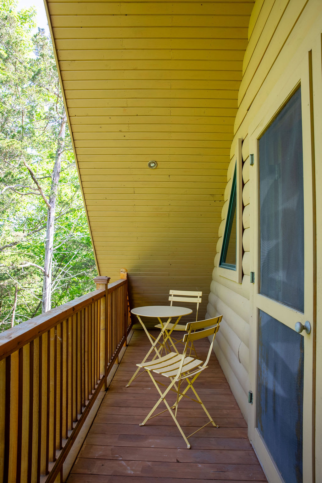 Narrow wooden balcony with a yellow slatted railing, featuring a small round table and two matching yellow folding chairs. Lush green trees are visible beyond the railing.