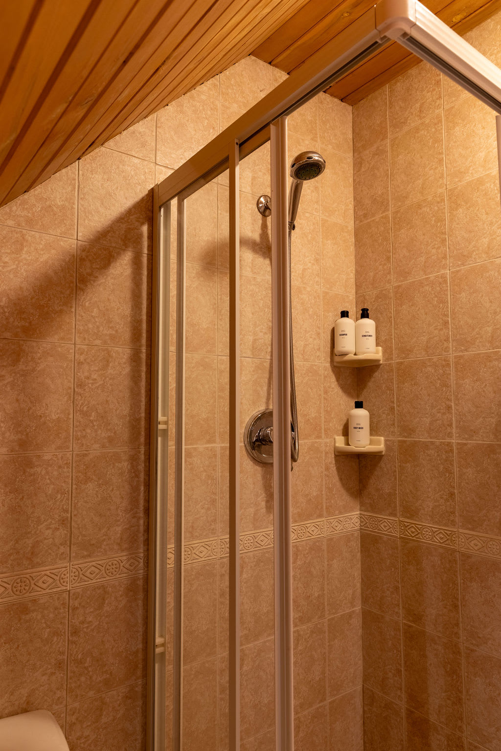 Tiled shower stall with light brown square tiles, a glass enclosure, and a silver shower head. Two white shelves hold toiletries, and the ceiling is slanted wood.