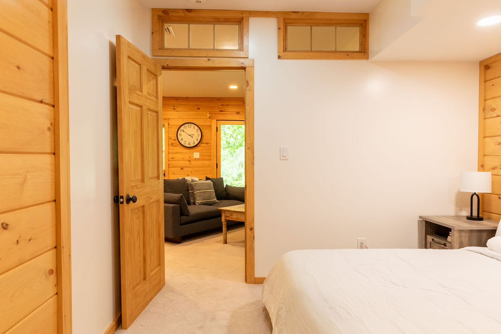 View from a bedroom with white walls and wood paneling, looking through an open doorway into a living area with a dark gray sofa and wood paneled walls. Transom windows are above the doorway.