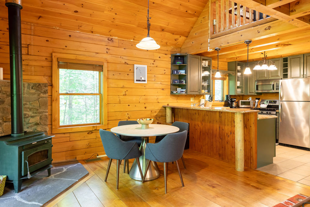 An open-concept dining area with light wood paneling and exposed beams, featuring a round white dining table with four grey chairs, adjacent to a modern wood-burning stove and a kitchen bar.