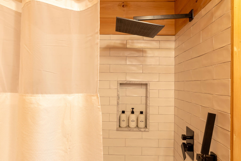 A modern shower featuring white subway tiles, a built-in niche with toiletries, and a black rain shower head, separated by a white shower curtain.