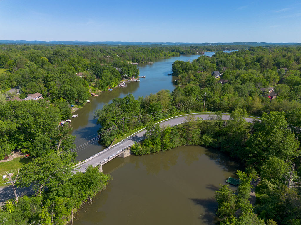 An aerial view of a winding river or lake surrounded by dense green forests under a clear blue sky, with a bridge crossing a narrow section of water. Several houses are visible along the shoreline.