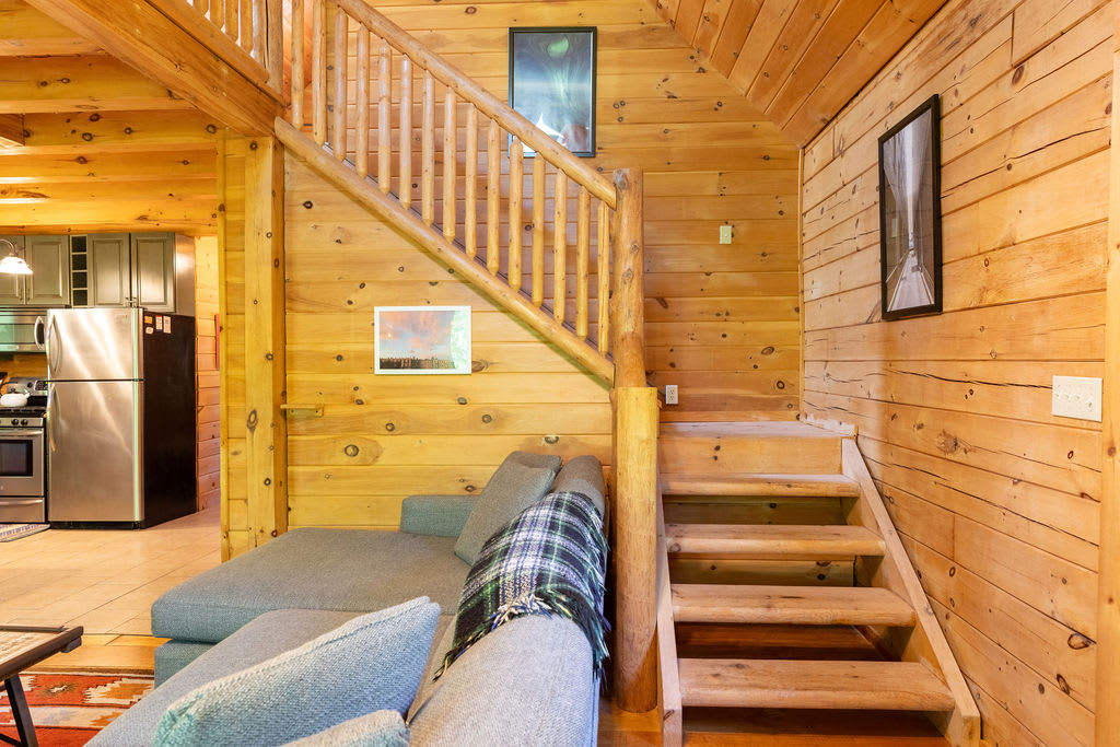 An inviting interior view showcasing a wooden staircase with log railings leading upstairs, a grey sectional sofa with a plaid throw, and a partial view of a kitchen in the background, all within a wood-paneled space.