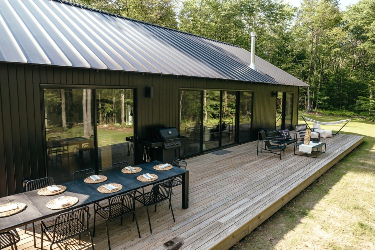A modern black cabin with a dark metal roof, featuring multiple large sliding glass doors opening onto an expansive wooden deck furnished with an outdoor dining table, grill, and lounge seating, all surrounded by lush green trees.