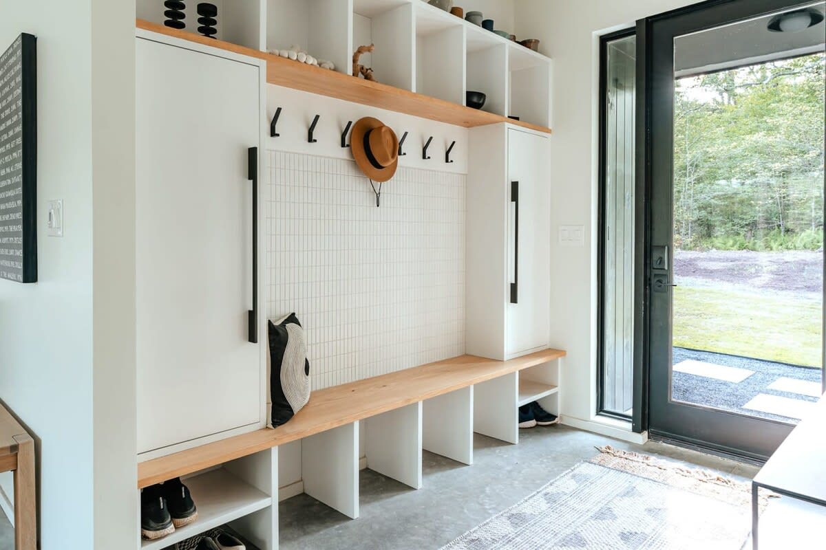 A modern mudroom with white built-in cabinetry, a light wood bench, and a white tiled accent wall with black hooks, offering shoe storage below and open shelving above, next to a black-framed glass door.
