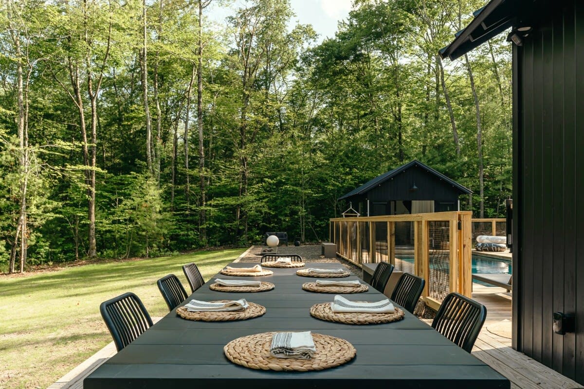 Expansive outdoor dining area on a wooden deck, featuring a long black table with multiple chairs and woven placemats, set against a lush green forest backdrop.