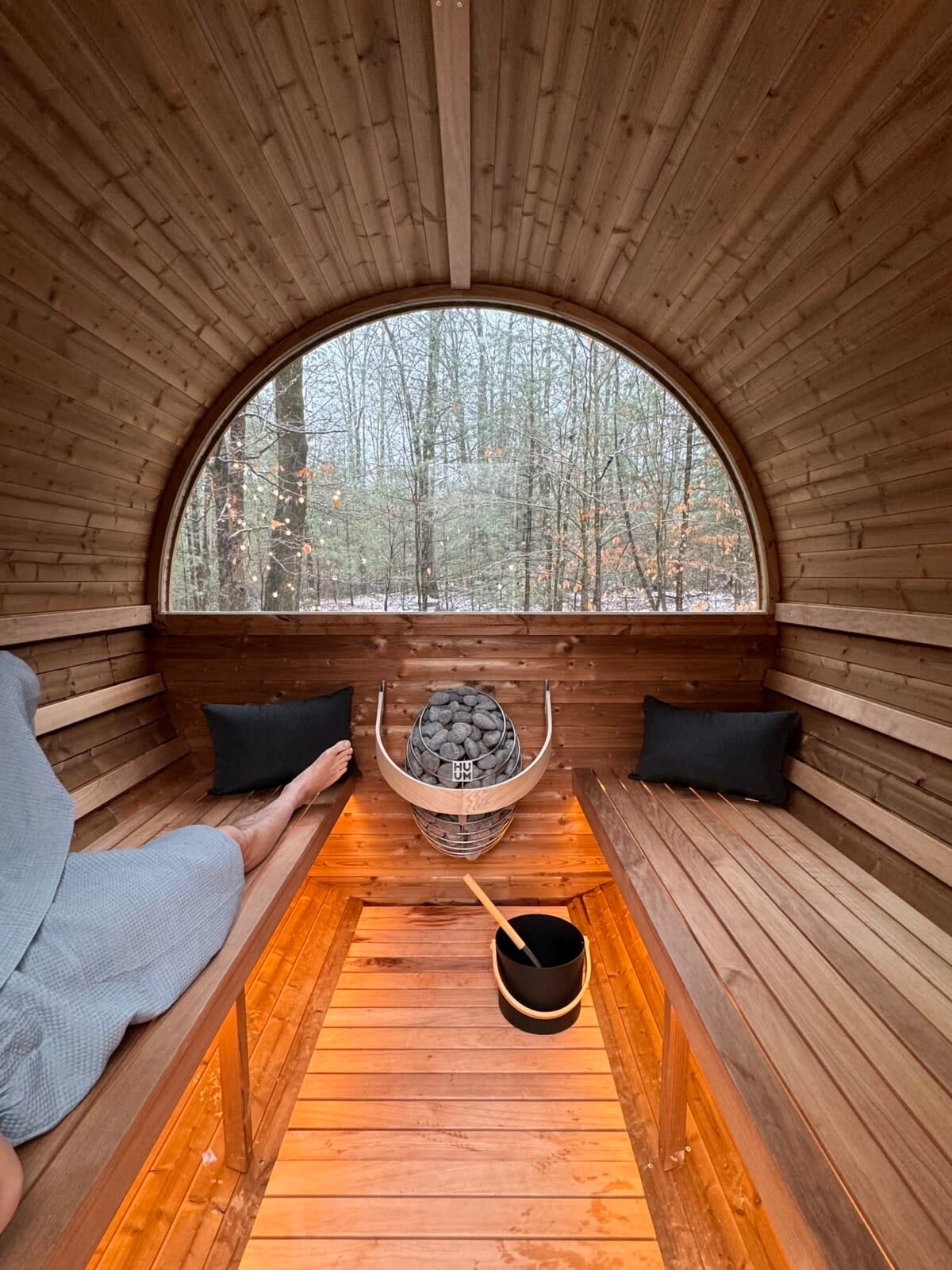 Cozy wooden barrel sauna interior with two benches, a stone heater, and a large arched window offering a view of a snowy forest.