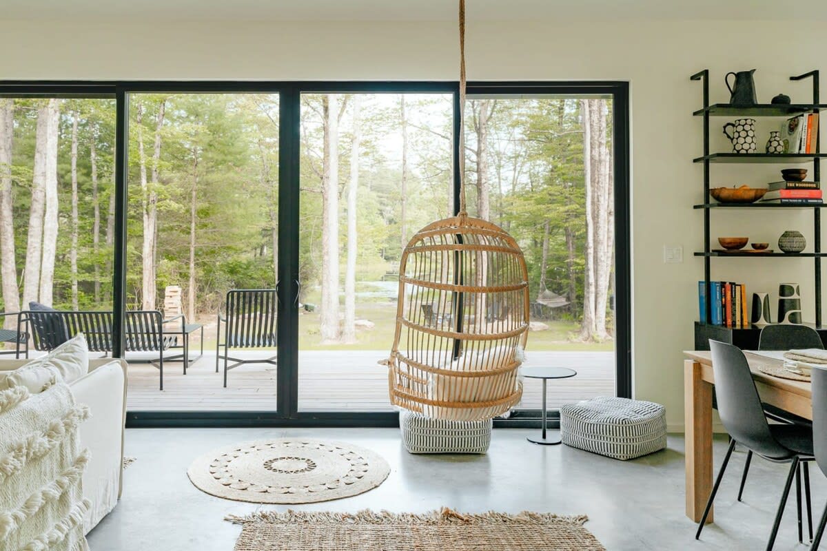 An open-concept living space with large sliding glass doors offering forest views, featuring a unique hanging wicker chair, a white sectional sofa, and a black shelving unit, with concrete floors and natural fiber rugs.