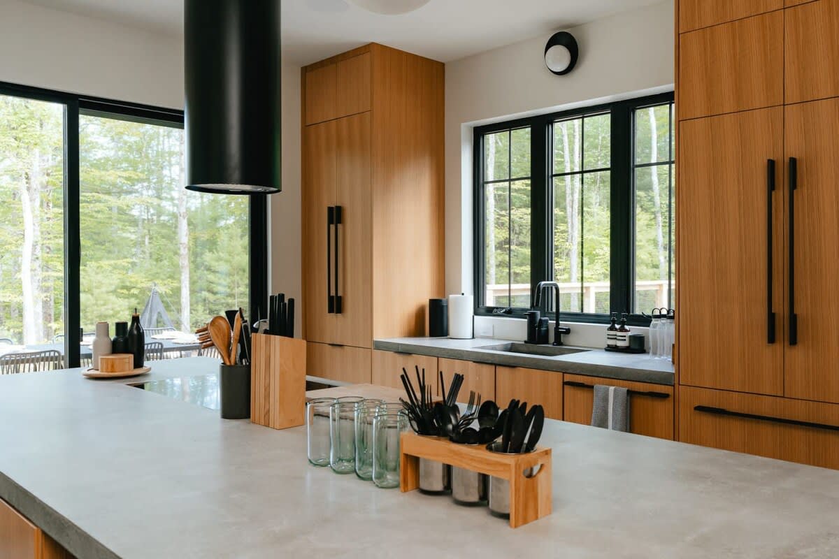 A modern kitchen with light wood flat-panel cabinetry, concrete countertops, and black-framed windows offering forest views, featuring a large island with a black cylindrical range hood and integrated appliances.