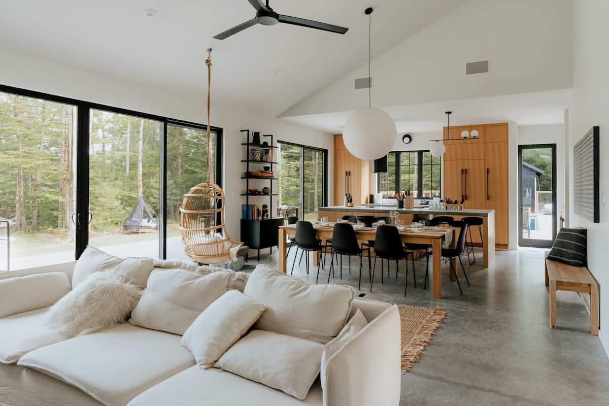A spacious open-concept living area with vaulted ceilings, connecting a white sectional sofa lounge, a dining area with a wooden table and black chairs, and a modern kitchen with light wood cabinetry, all overlooking a forest through large glass doors.