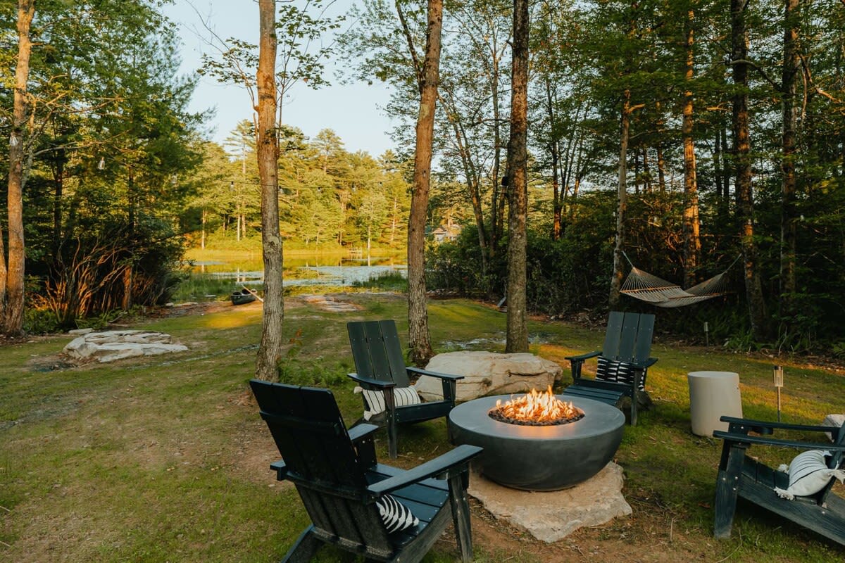 A circular fire pit with a concrete base is surrounded by four black Adirondack chairs on a grassy lawn, with tall trees and a glimpse of a lake in the background under a golden sky. A hammock is also visible.