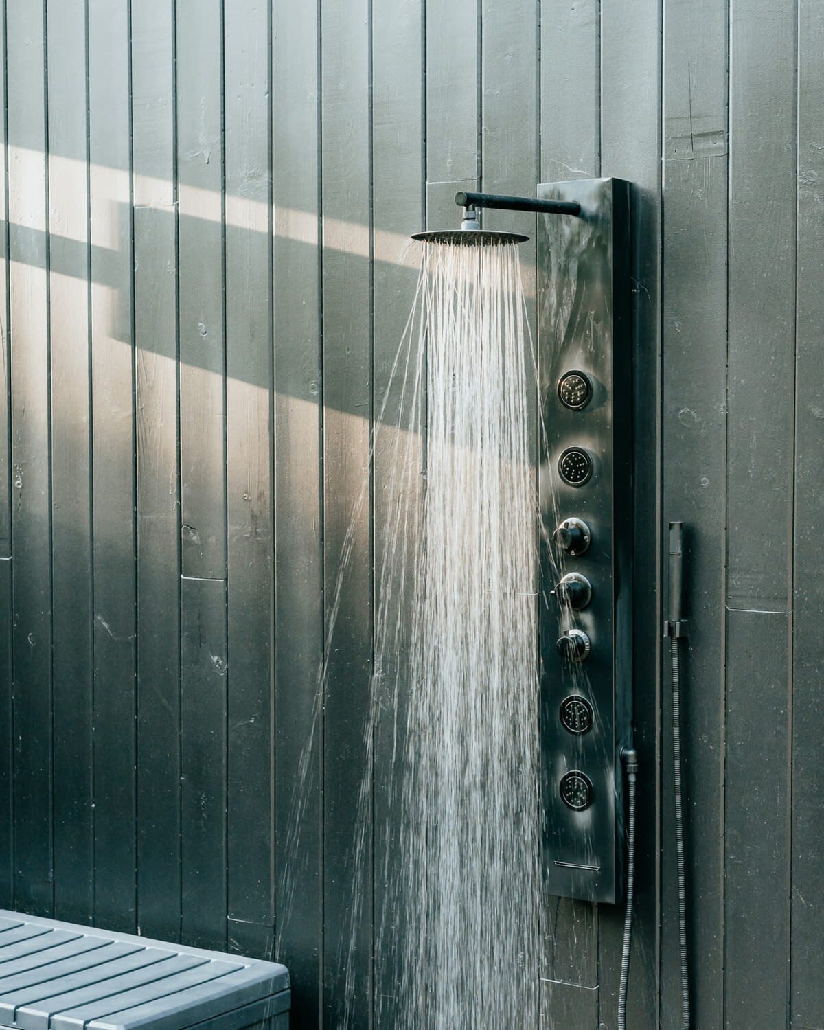 A close-up of a modern outdoor shower with multiple jets and a rainfall showerhead, spraying water against a dark wooden slatted wall. A light-colored bench is visible at the bottom, suggesting a changing or seating area.