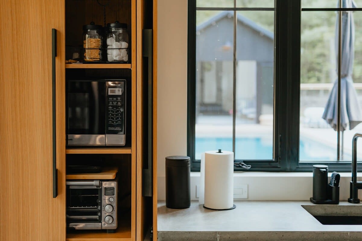 Built-in light wood cabinetry houses a microwave and a toaster oven, with a concrete countertop and a large black-framed window offering a view of a pool and outdoor area.