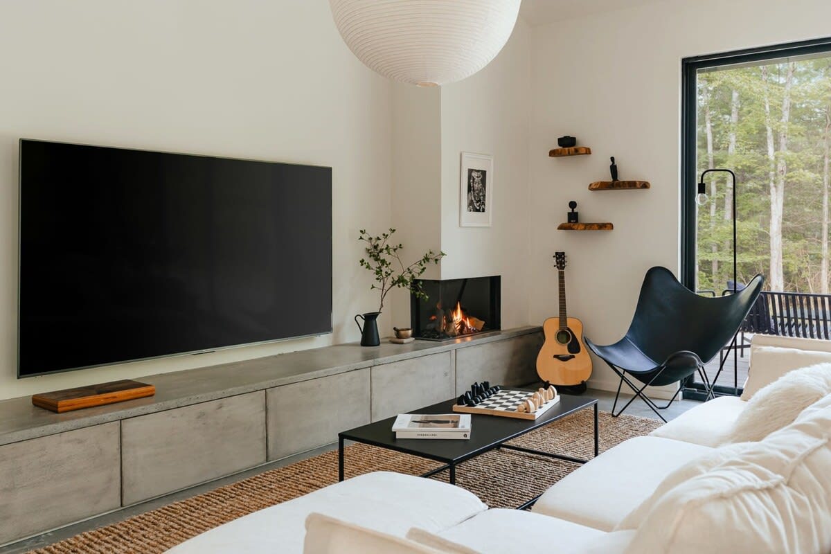 A contemporary living room featuring a large wall-mounted TV above a concrete media console, a modern fireplace, a black coffee table with a chess set, and a white sectional sofa, all set on a textured jute rug.