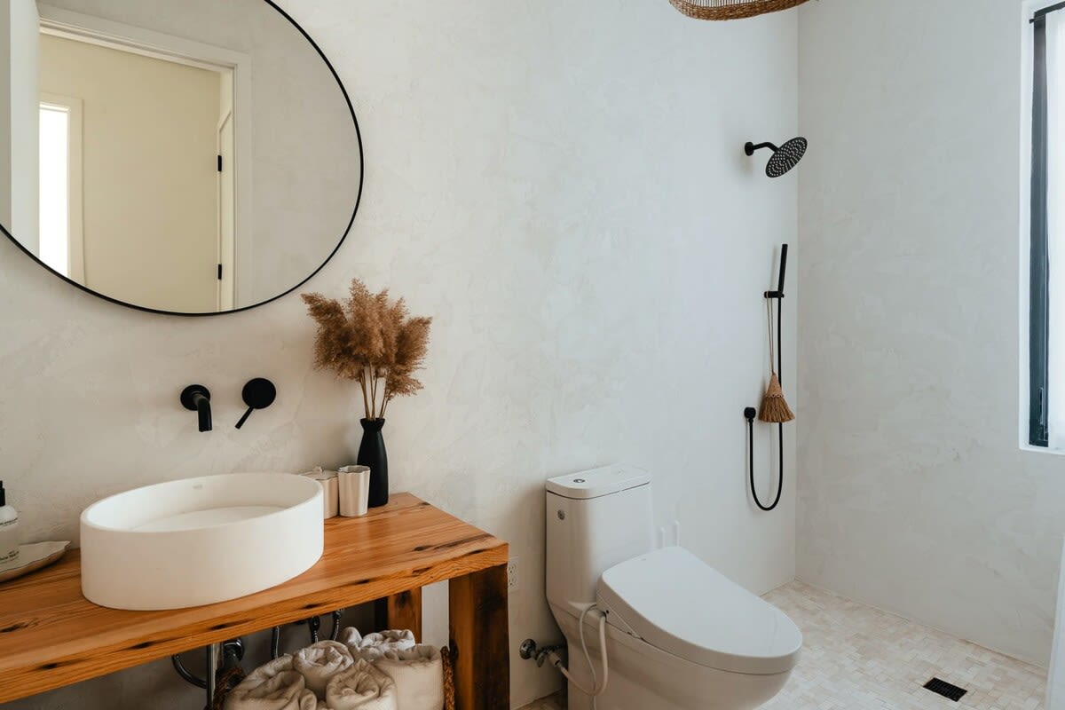 Modern bathroom with textured white walls, a large round mirror, a white vessel sink on a rustic wooden vanity, and black matte fixtures. A toilet and shower with black hardware are also visible.