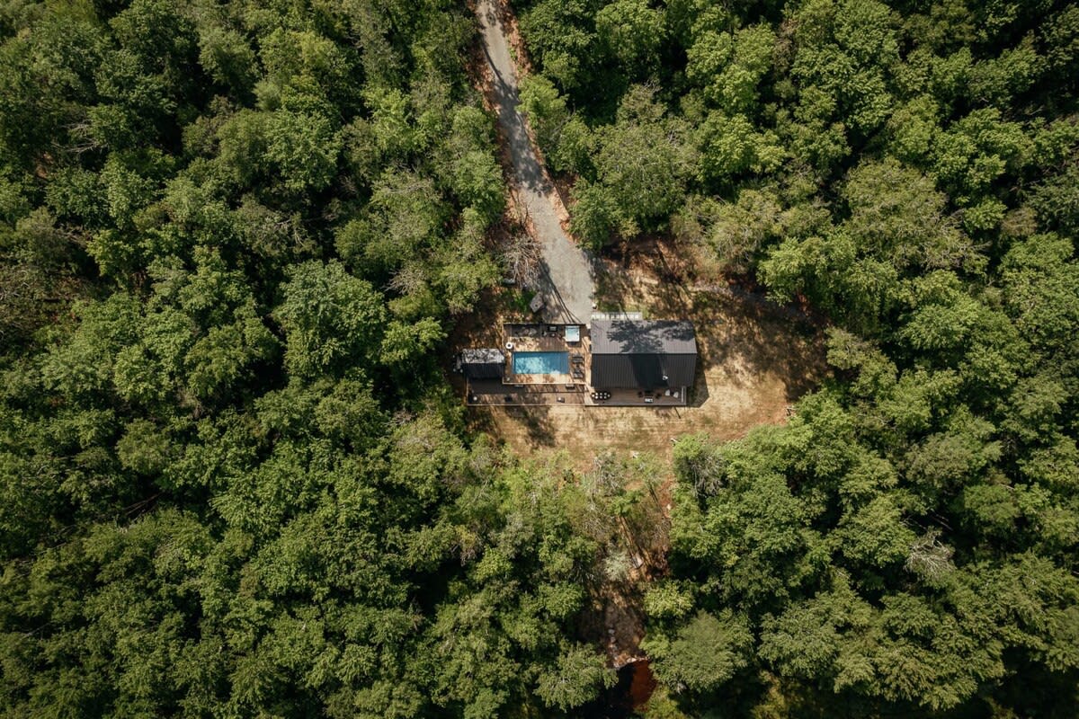 An aerial view of a black modern cabin with a dark roof, nestled in a dense green forest. A rectangular swimming pool and wooden deck are visible next to the cabin, with a gravel driveway leading to the property.